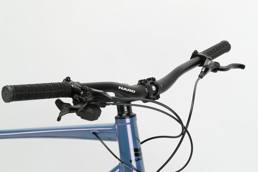 Close-up of a Haro MTB Beasley 27.5 DLXs metallic blue aluminum frame, black handlebars with textured grips, brake levers, and gear shifters. Cables run visibly from the handlebars against a plain light background.