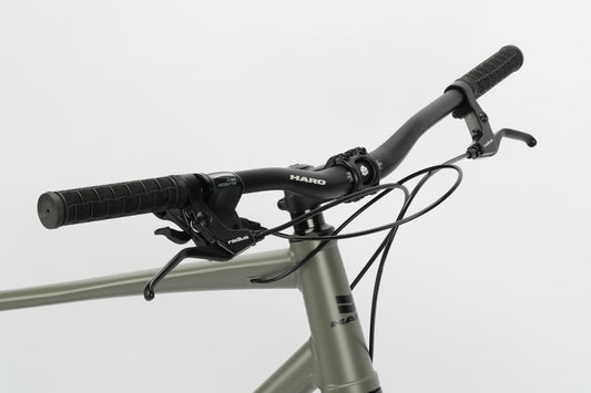 The Beasley 27.5 by Haro MTB features a close-up of its matte olive green handlebar with grips, brake levers, gear shifters, and mechanical disc brakes on a lightweight aluminum frame. The black cables are neatly routed against a white background with HARO branding.