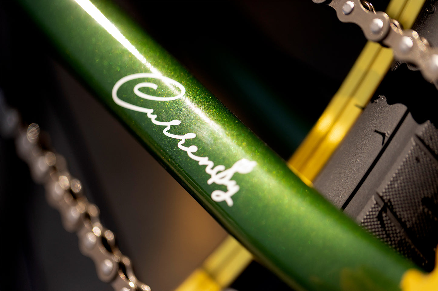 Close-up of a green bicycle frame with Curency in white script, evoking the stylish Haro Bikes Jetlife x CEEK 29”. Accented by bright lighting and surrounded by bike chains, it’s a must-see for any B.M.F. big bike enthusiast.
