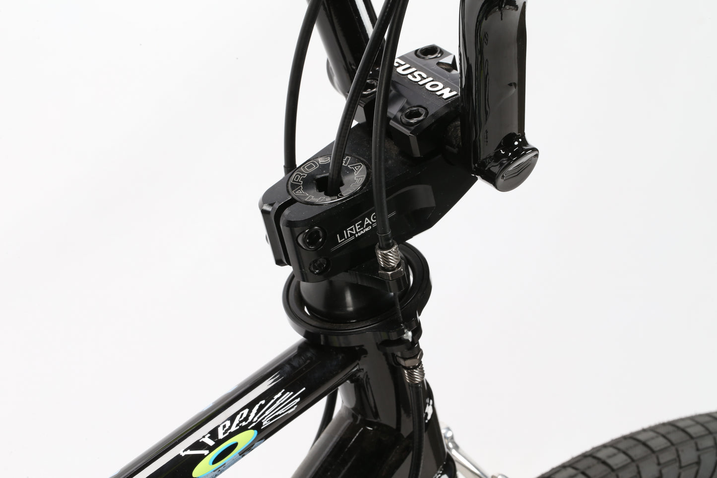 Close-up of the black handlebar and stem on the Haro Bikes Lineage Sport Bashguard 26 2023 BMX, showcasing brand logos and cables. The bikes chromoly frame sports a vibrant multicolored design enhancing both its durability and sleek build.