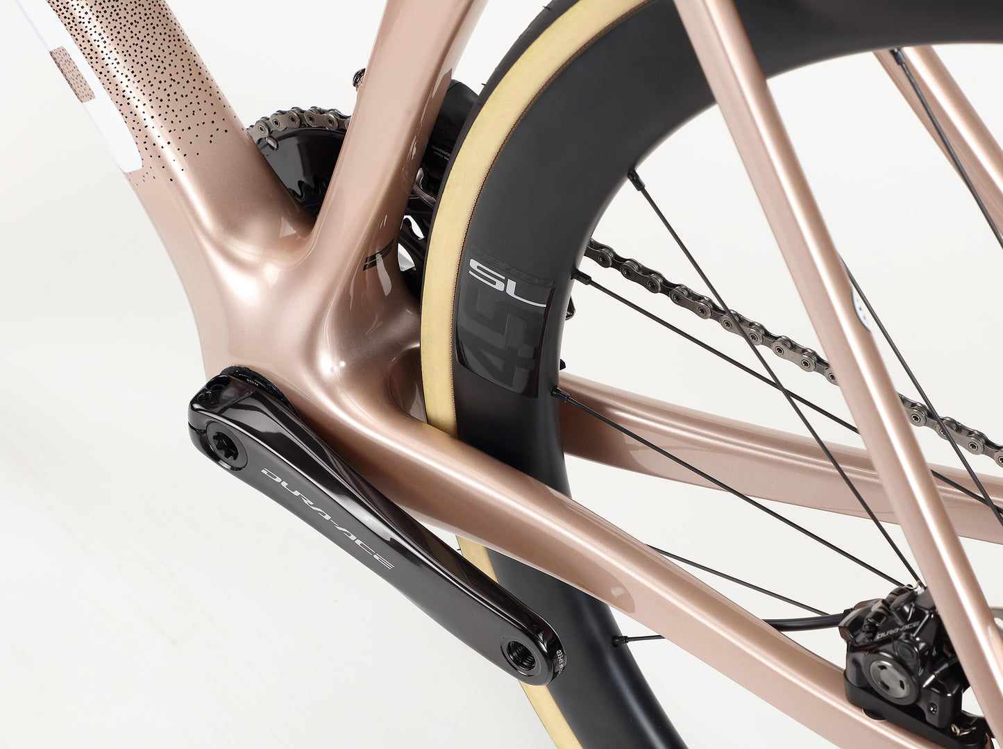 Close-up of the Rivette Carbon Top Shelfs rear wheel and drivetrain from Haro Bikes, featuring aero tube shapes and a metallic rose gold frame. The black tire with a cream edge is complemented by the black Dura-Ace crank, set against a plain white background.