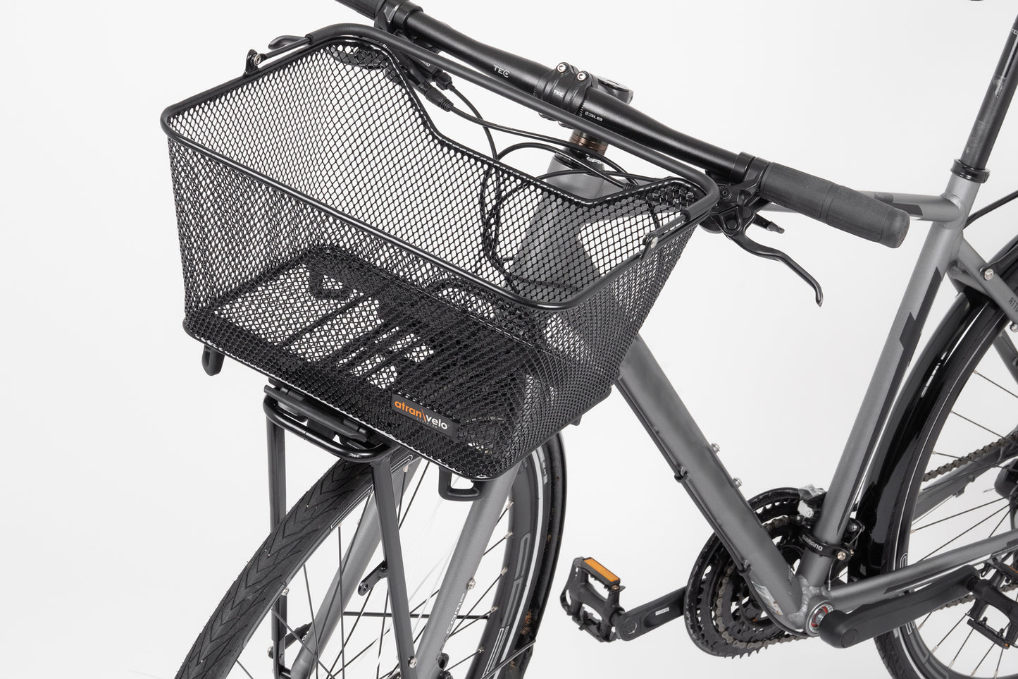 A close-up of a bicycle features Atran Velos Daily Basket Small, a durable steel basket on the front. The bike has a gray frame with black grips on the handlebars, while the background is softly blurred.