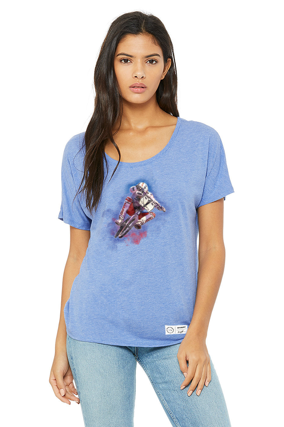 A woman with long dark hair wears the Haro Bikes Windy Osborn Womens Tuck Slouchy Shirt. The blue tee, featuring a motocross rider design, boasts poly viscose fabrication and a wide scoop neckline. It pairs effortlessly with her light blue jeans as she stands against a plain white background.