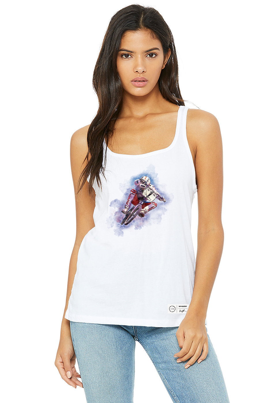 The woman models a white Windy Osborn Womens Tuck Tank by Haro Bikes, featuring a motorcyclist on the front. Her long dark hair flows over light blue jeans against a white backdrop. This tank, made from Airlume combed cotton, offers both style and comfort.