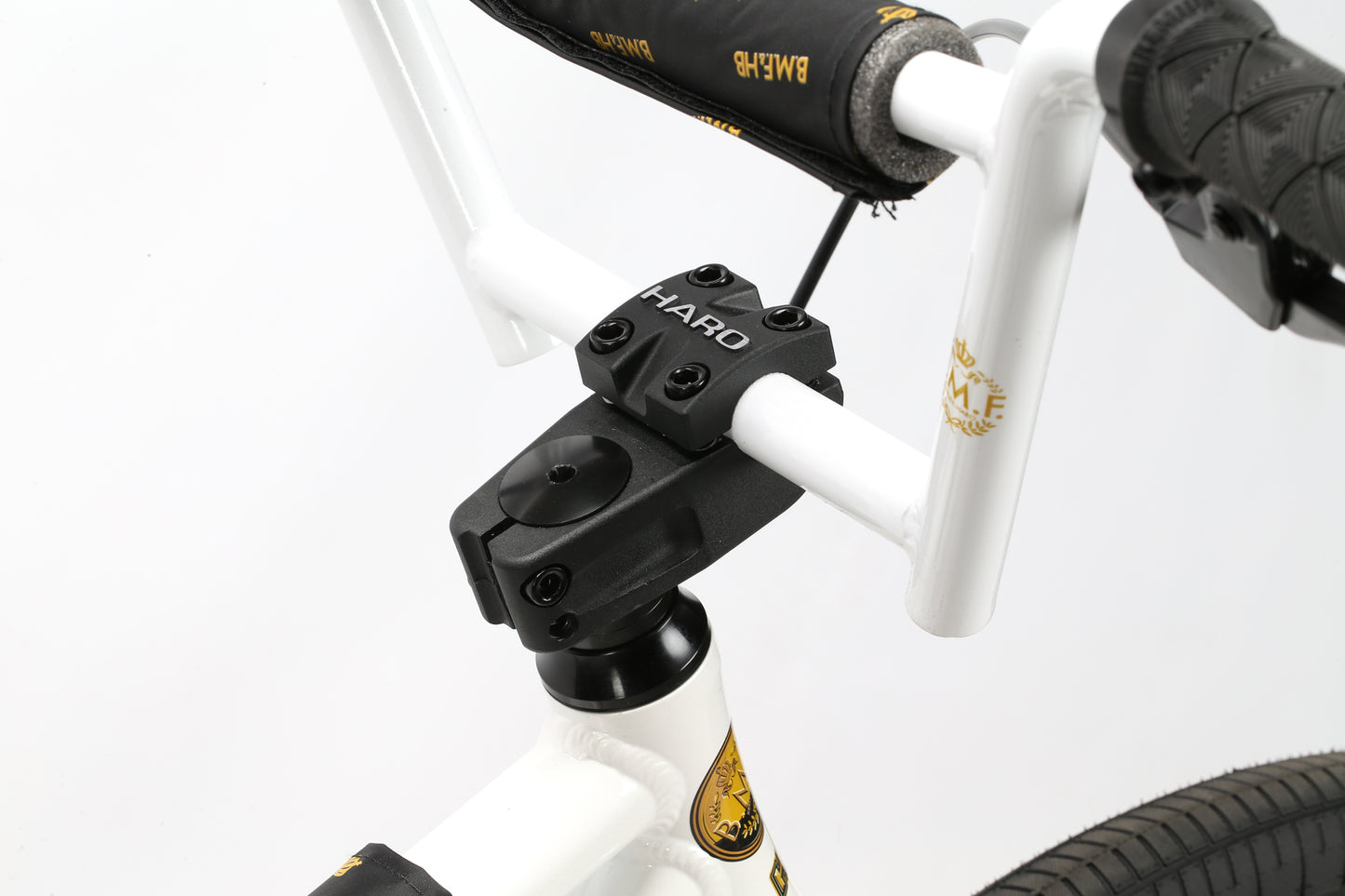 Close-up of the Haro Bikes B.M.F 29s white handlebars, with a black stem and textured grips. Gold accents highlight the frame, including a small emblem. The plain white background emphasizes Bike Lifes essence.