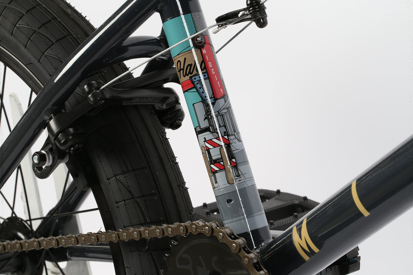A close-up of the Haro Bikes Mid City BMX reveals a vibrant abstract cityscape design on the seat tube. The image highlights the bikes chain, tire, and pedal in a black and teal color scheme on its resilient chromoly frame.