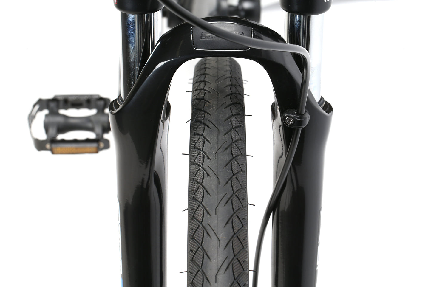 Close-up of the Westlake MicroShift by Haro MTBs front fork and wheel. The black fork encloses a treaded black tire with an attached brake cable. A pedal is partially visible on the left against a plain white background.