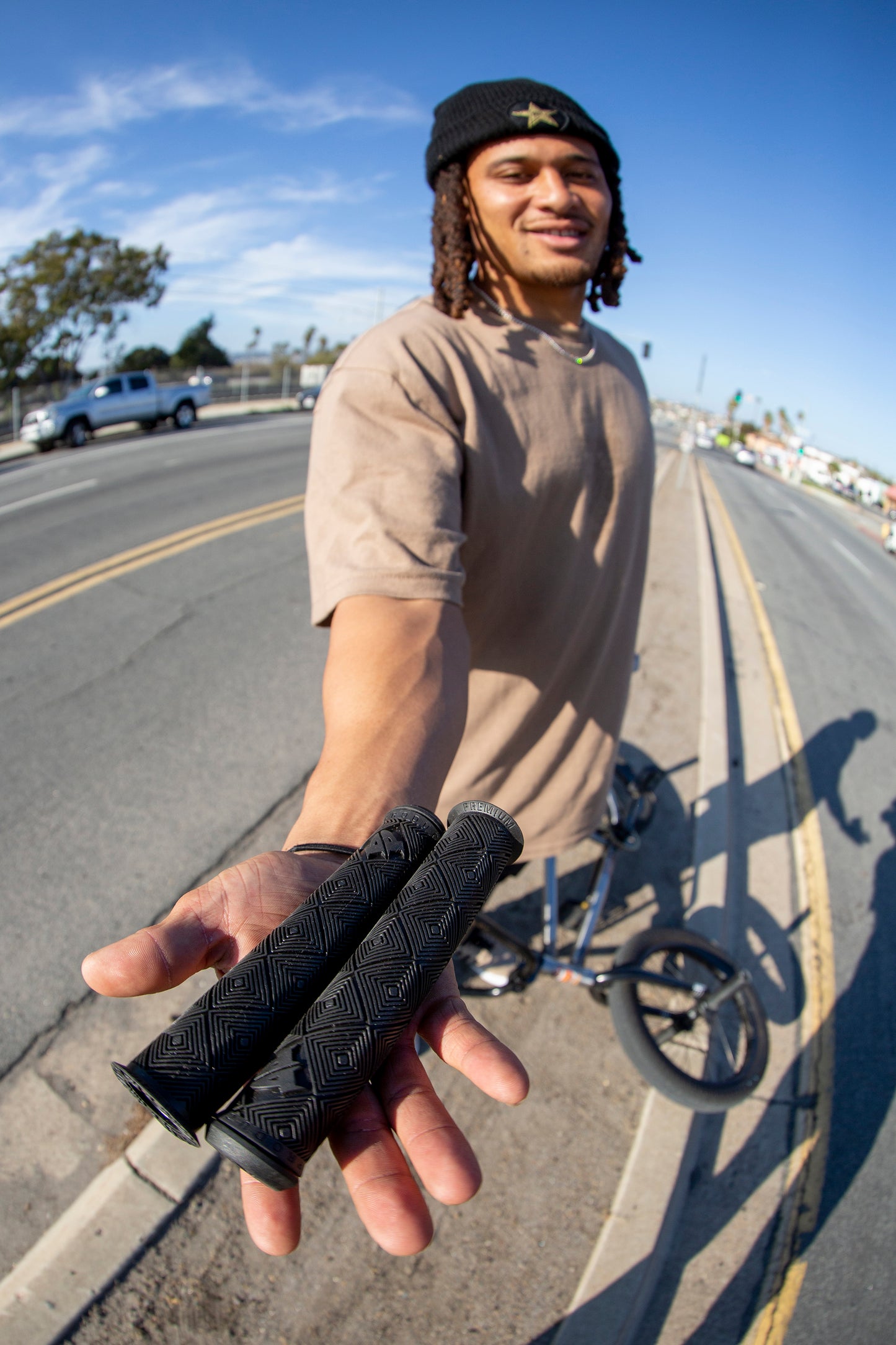 A person on the street holds out two CK Grips from Premium. A bicycle is beside them, and the road extends into the distance. They wear a beanie and tan shirt under clear blue skies.