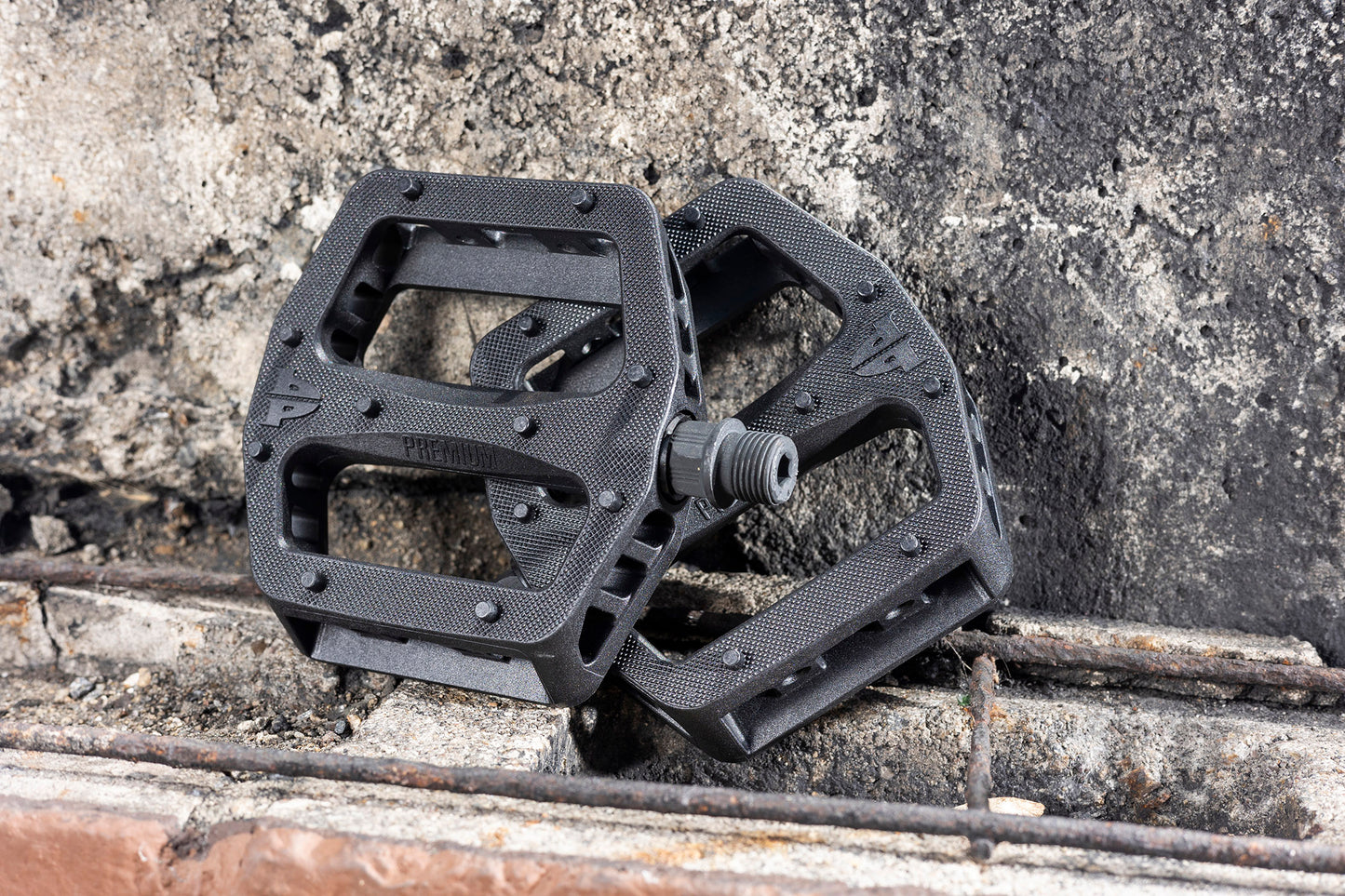 Close-up of two Premium Plastic Kinetic Pedals with textured surfaces against a rough, gray concrete background. The lightweight nylon pedals are crisscrossed, showcasing their angular and open-frame design for maximum traction.