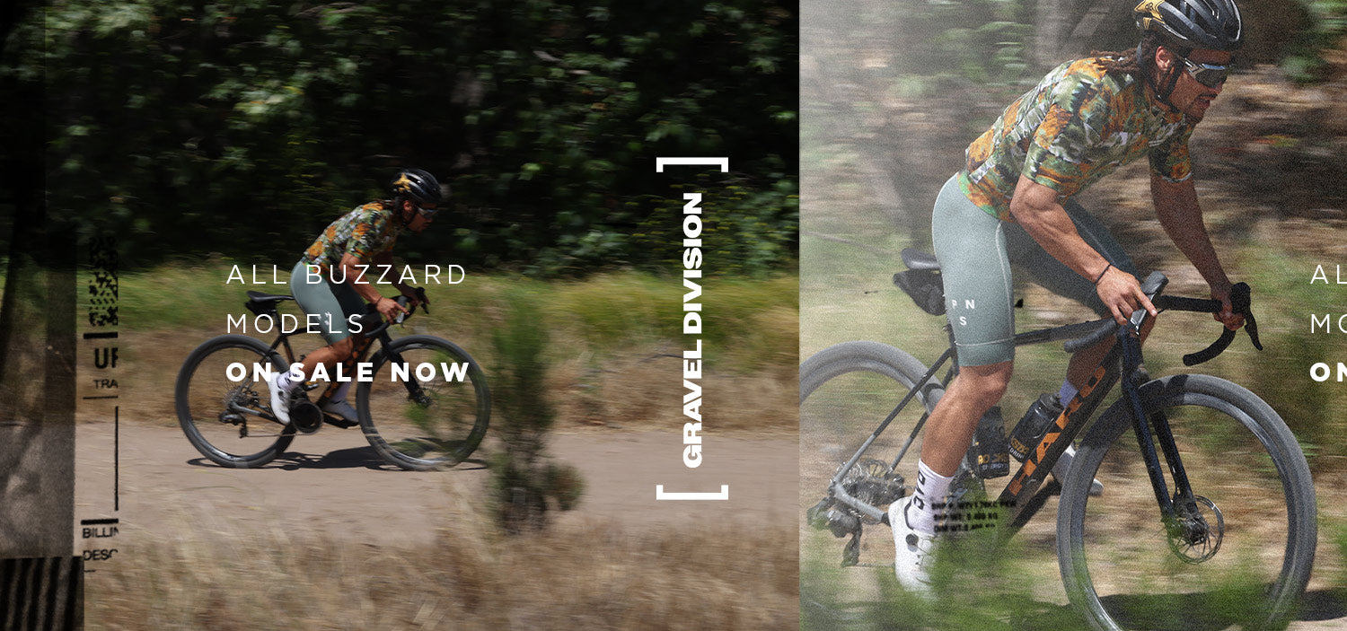 A cyclist in black gear rides a gravel bike on a sandy trail through a dry, bushy landscape near a lake and mountains.