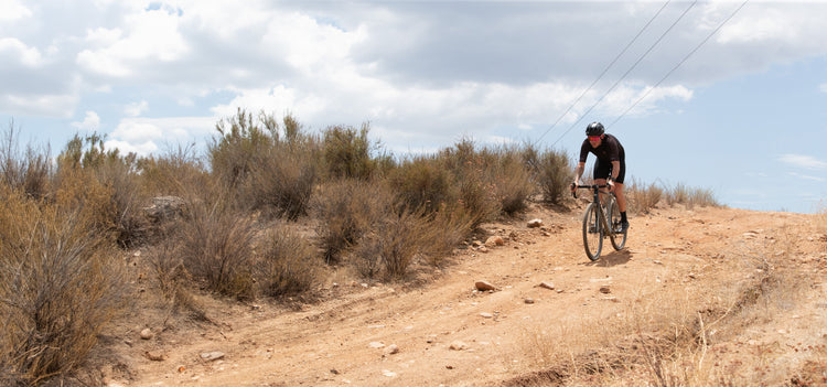 A cyclist in black gear rides a gravel bike on a sandy trail through a dry, bushy landscape near a lake and mountains.