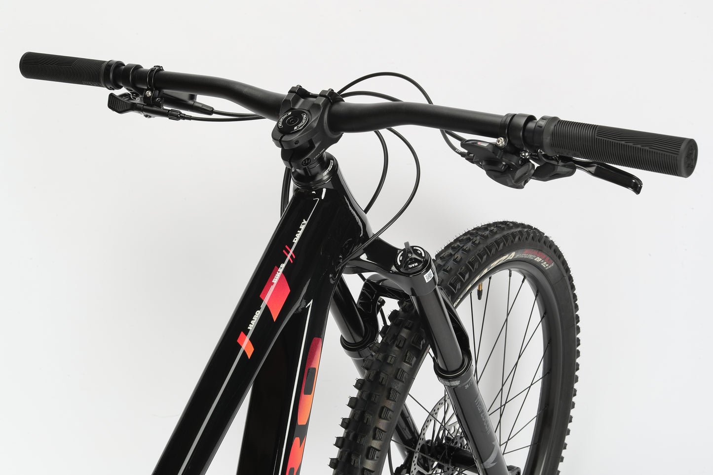 Close-up of the black handlebar and front wheel of a Daley Alloy 2 all-mountain 29er by Haro MTB, with an aluminum frame featuring red and white detailing, rugged off-road tires, and set against a plain white studio backdrop.