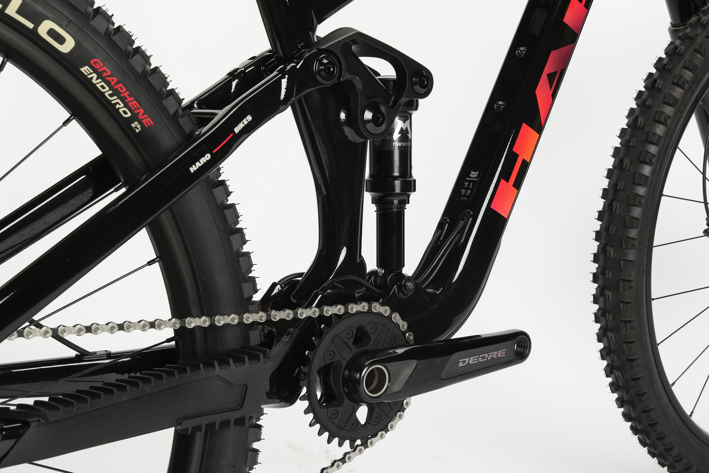 The close-up image of a black all-mountain 29er showcases the drivetrain and suspension system. It highlights the chain, gears, aluminum frame elements, with Deore on the crank arm and Hard branding on the frame. This Haro MTB Daley Alloy 2 is ideal for conquering any terrain.