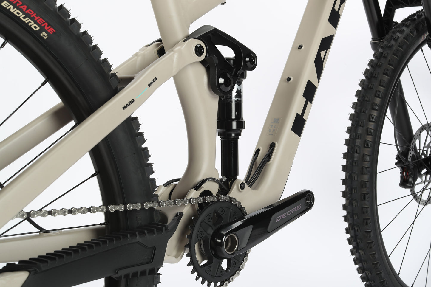 Close-up of the Daley Alloy 2s suspension and drivetrain on this beige 29er, highlighting chain, crankset, and tire treads. The rugged design of Haro MTBs all-mountain aluminum frame makes it ideal for challenging terrains.