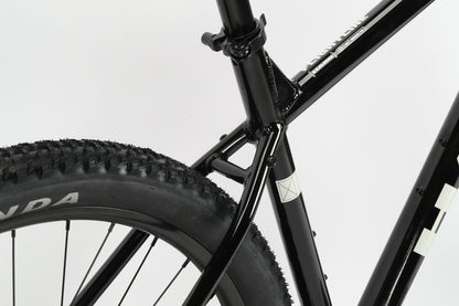 Close-up of a Haro MTB Flightline LTD 29 featuring the rear wheel, frame, and seat tube. The tire tread is prominent, with a small white geometric decal on the frame. Equipped with Shimano Cues for smooth shifting, the backdrop is plain and minimalistic.