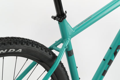 Close-up of the teal frame of Haro MTBs Flightline LTD 29 highlights the rear wheel with a textured tire, Shimano Cues, seat post, and Tektro hydraulic brakes against a plain white background.