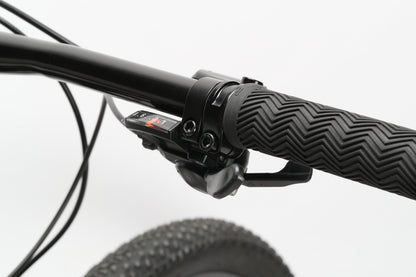 Close-up of the Haro MTB Flightline 1 29s handlebar with a textured black grip and Shimano 8-speed Essa gear shifter, featuring a small display for gear settings. The tire and hydraulic disc brakes hint at its hardtail 29er design.