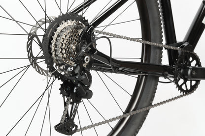 Close-up of the rear wheel on a Haro MTB Flightline 1 29 bicycle, highlighting the Shimano 8-speed Essa derailleur, chain, and gears. The metallic and black parts display the detailed mechanics of the drivetrain against a white background.