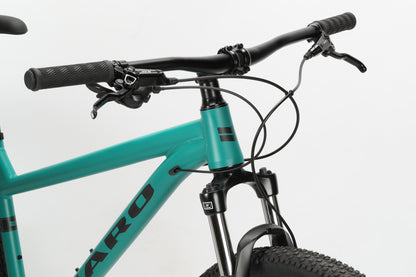 Close-up of the front section of a Haro MTB Flightline 1 29 hardtail mountain bike in teal, highlighting the handlebars, Shimano Essa 8-speed brake levers, cables, and part of the front suspension fork. The bike frame displays bold black lettering against a plain white background.