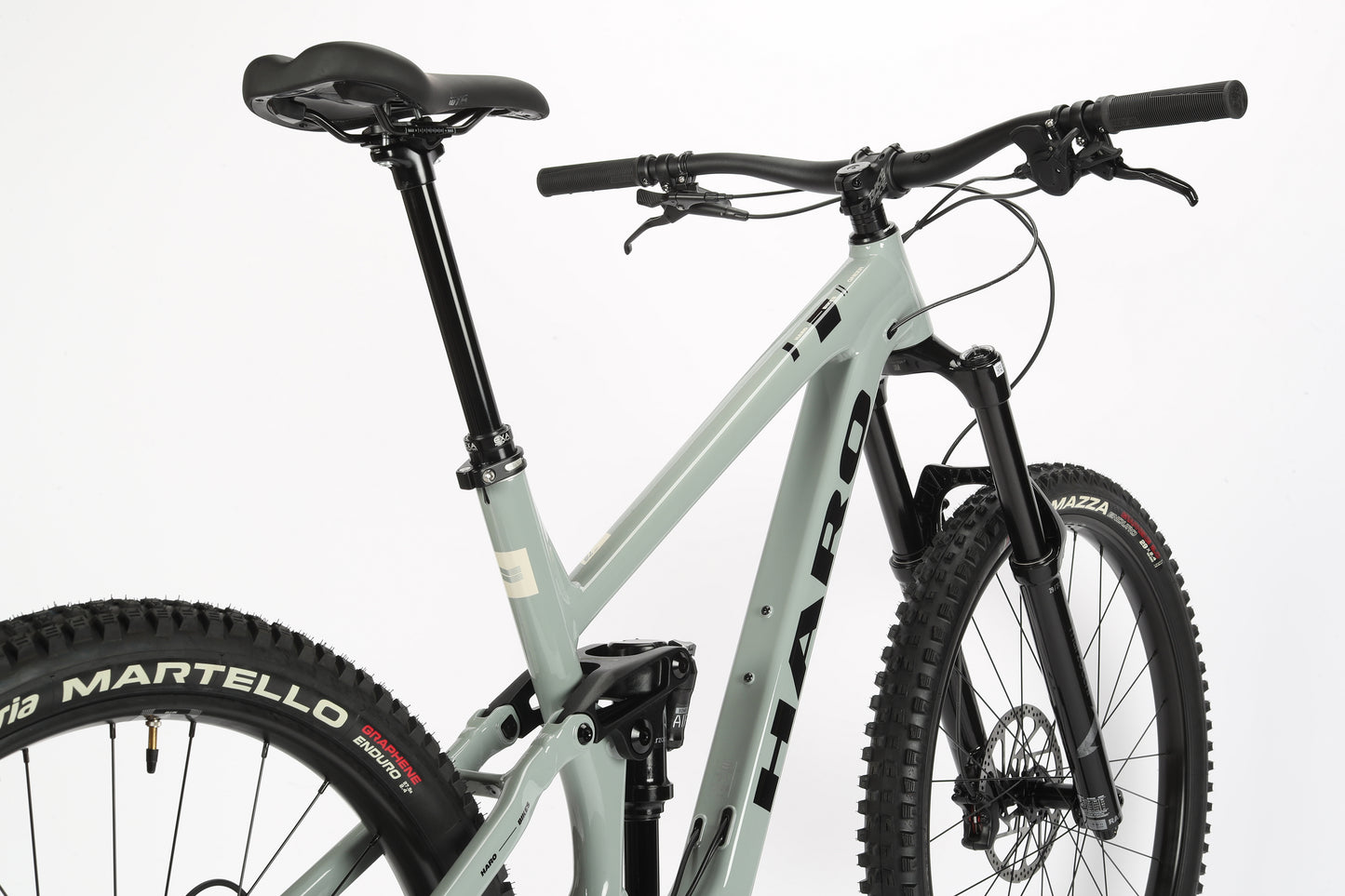 A close-up of the Haro MTB Greer Alloy 2 mountain bike highlights its light gray performance alloy frame, black handlebars, and seat. It features thick Vittoria Martello tires, a suspension system, a mullet wheel configuration, and HARO branding on the frame.