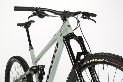 Close-up of a light gray Haro MTB Greer Alloy 2 mountain bike with black handlebars, featuring a performance alloy frame and front suspension fork. Its prominent tire is branded Vittoria Mazza, and the mullet wheel configuration complements the sloping top tube against a white background.