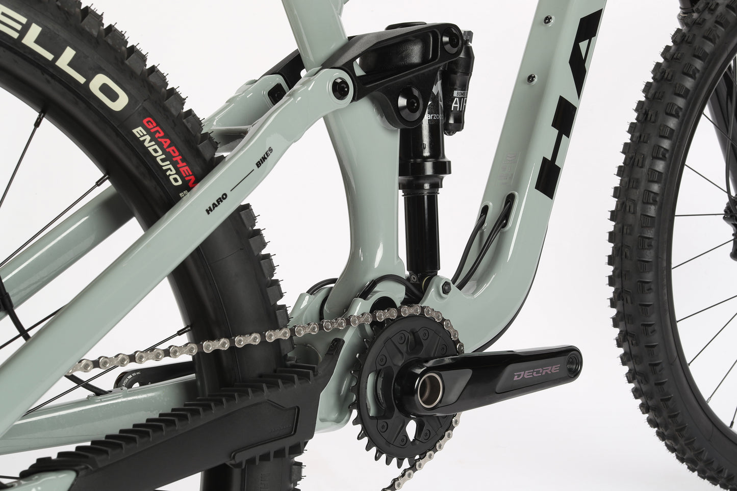 A close-up of the Greer Alloy 2s drivetrain highlights a rear suspension, chain, and Gripper Enduro tire. It features a light gray performance alloy frame and a crank arm labeled Deore. This Haro MTB offers a sleek design perfect for adventure seekers.