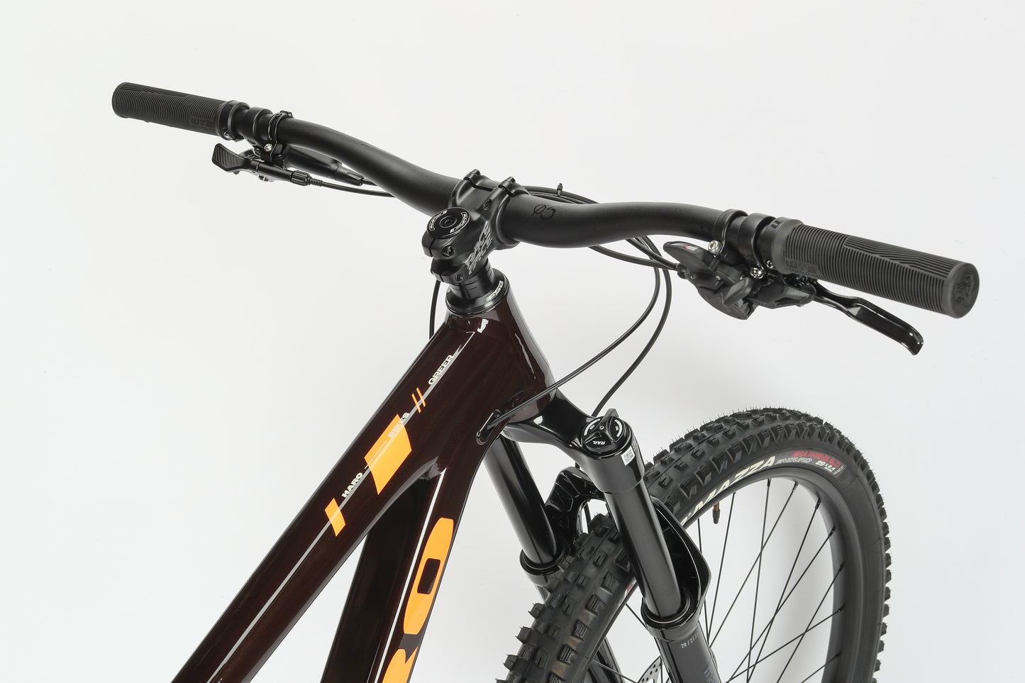 Close-up of a Haro MTB Greer Alloy 2, featuring black handlebars, brake levers, and suspension fork parts. The dark performance alloy frame with orange accents and the thick tire hint at its mullet wheel setup. Background is white.