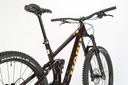 A close-up of a dark brown Haro MTB Greer Alloy 2 mountain bike shows its thick tires, performance alloy frame, and disc brakes. The sleek design is highlighted by Haro Greer in orange on the frame and handlebars against a plain white background.