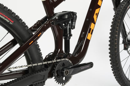 Close-up of the Greer Alloy 2s rear suspension, highlighting its performance alloy frame, shock absorber, drivetrain with chainring and pedals. The Haro MTB bike features chunky off-road tires and orange brand lettering on the frame for ultimate trail adventures.