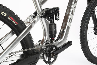 Close-up of a silver mountain bikes rear suspension system and drivetrain, featuring a chain, Fox suspension, and tire. The aluminum frame showcases a modern design with visible Haro MTB branding and intricate components. Product: Greer Alloy LTD 2.