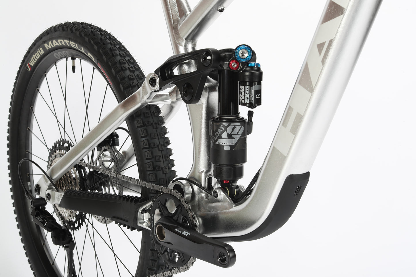 Close-up of a mountain bikes rear suspension with Fox suspension. The silver aluminum frame, branded Vitus, highlights a shock absorber and intricate linkage, showing the textured tire and crankset similar to Haro MTBs Greer Alloy LTD 2 model.