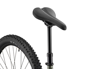 Close-up of Double Peak’s black seat and dropper post, with rear wheel and tire partially visible against a white background.