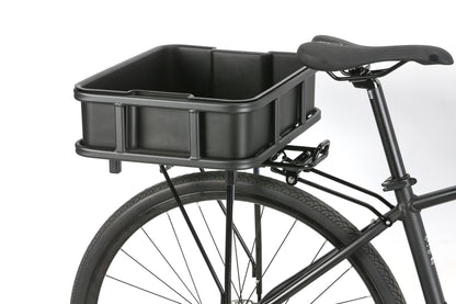 A close-up of a black bicycle emphasizes the rear section featuring Atran Velos Bakery Crate, a large rectangular cargo basket over the back wheel. The saddle is visible, complementing the black frame and tire prominently displayed.