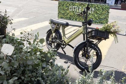 An Atran Velo Bakery Crate electric bike with a sturdy, cargo-style design is parked near a rack. Its front basket holds colorful flowers. In the background, a Little Italy sign is visible amid surrounding greenery and plants.