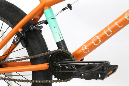 Close-up of the Boulevard 20.5s vibrant orange frame, showcasing its chain, pedals, and rear wheel. A turquoise section with a black graphic displays Haro Bikes brand name in partially visible white lettering, promising a long-lasting ride for BMX enthusiasts.