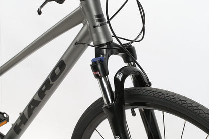 Close-up of a bicycles front section, showing the handlebars, brake cables, and front suspension fork. The metallic gray frame features Haro MTB branding for the Bridgeport ST MicroShift model. The textured tread pattern on the black front tire is also visible.