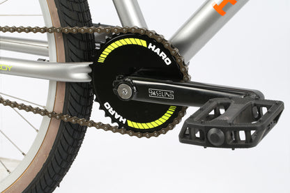 Close-up of the DMC 24s drive train with silver frame and black crank arm labeled Haro Baseline. The 24 wheels have a black and neon yellow chainring. This Haro Bikes BMX features textured black pedals, wide tread tires with brown sidewalls, and Freestyler bars for ultimate control.