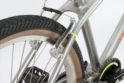 Close-up of the Haro Bikes DMC 24 BMX frame highlighting the rear wheel, tire, brake, and part of the pedal. The silver metallic frame shows visible welds. It features 24 wheels with black tread and beige sidewalls, contrasting stylishly with its rugged design.