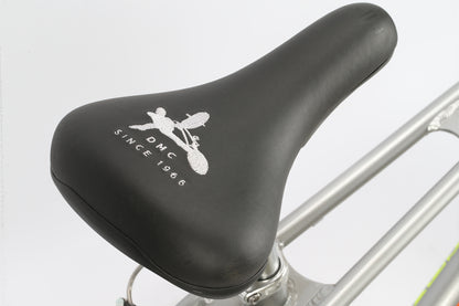 A close-up of the black DMC 24 bicycle seat from Haro Bikes, featuring a cyclist logo and DMC Since 1966 in white, pairs perfectly with freestyler bars. Its attached to a silver frame designed for BMX track agility.