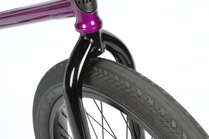 Close-up of a Haro Bikes Danas front wheel and fork. The glossy black fork complements the metallic purple chromoly frame. Equipped with Premium CK tires featuring a unique tread pattern, this setup ensures responsive geometry against a crisp white background.