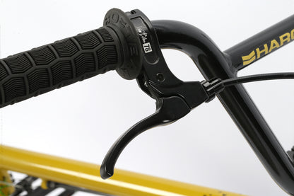 Close-up of a Leucadia black handlebar by Haro Bikes, featuring a textured grip with a logo and the letter B. A brake lever is visible, reflecting the sleek design typical of skateparks, with part of the yellow frame in the background.