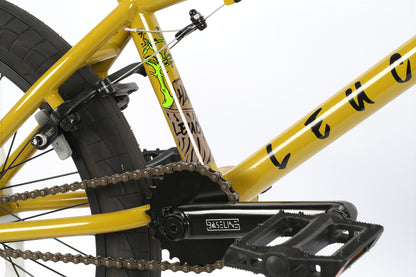 Close-up of a yellow bicycle frame by Haro Bikes, featuring their Leucadia model amidst a lively neighborhood. The image showcases the gears, chain, and rear wheel alongside a decorative sticker on the frame, capturing a day filled with skateboards and outdoor activities.