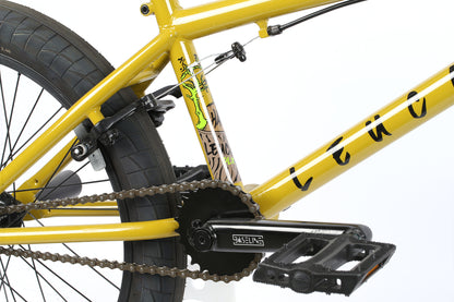 Close-up of the Leucadia DLX yellow freestyle BMX bike by Haro Bikes showcases the chromoly cranks, pedals, and part of the frame. It features a chunky rear tire, chain, and unique graphic designs near the seat tube against a white background.