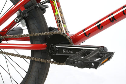 Close-up of the Leucadia DLX by Haro Bikes: its chromoly cranks gleam against a red frame with black details. The black pedal pairs well with the thick, textured tire, while a colorful sticker adds flair. Adding a gyro detangler would complete this freestyle BMX bike for tricks.